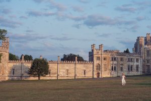 A woman in a white dress walks across a grassy field in front of a large, historic stone castle with towers and battlements under a partly cloudy sky.