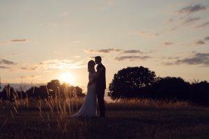 A bride and groom embrace at sunset in a field, silhouetted against the sky with tall grass and trees in the background.