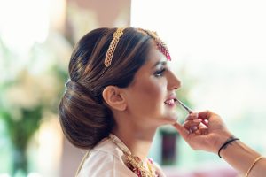 A woman in traditional attire with a jewelled headpiece is having her make-up done, as a hand applies lipstick to her lips. She is smiling and looking ahead, with a soft, blurred background behind her.