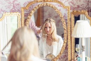 A woman with long blonde hair sits in front of an ornate gold mirror, having her hair styled. She wears a white dressing gown and looks at her reflection. A glass of champagne and a white lamp are on the table beside her.