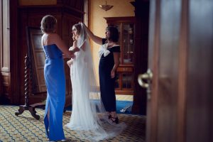 A bride in a white dress and veil stands with two women in elegant dresses, one adjusting her veil, in a warmly lit, wood-panelled room with patterned carpet and vintage furnishings.