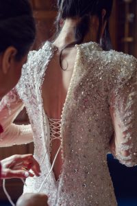 A person in an ornate, beaded wedding dress is being helped by another person to lace up the back of the gown. The image is focused on the intricate details of the dress and the hands adjusting the laces.