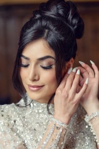 A woman with dark hair styled in an elegant updo wears a sparkling, sequinned outfit and delicate make-up. She smiles softly with closed eyes whilst adjusting an earring.