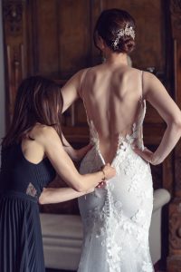 A woman helps another woman zip up the back of her lace wedding dress, revealing delicate floral details and an elegant updo hairstyle adorned with a jewelled hairpiece.