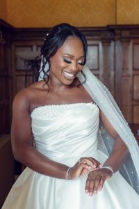 A smiling bride in a strapless white wedding dress and veil looks down at her hands, showing off her ring and bracelet, in a warmly lit, elegant room with wood panelling.