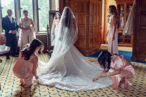 A bride in a sparkling gown stands facing a mirror, surrounded by bridesmaids in pink dresses adjusting her train, whilst family members watch and smile in a sunlit, wood-panelled room.