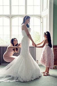 A bride in a white wedding dress stands holding hands with a young girl in a pink dress, while another woman kneels, adjusting the bride's dress, in front of large windows with natural light streaming in.