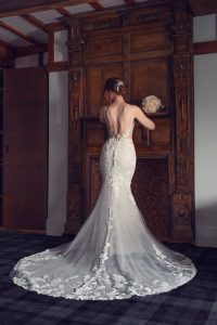 A bride in a fitted, lace-back wedding dress stands facing a wooden mantelpiece, her hair styled in an elegant updo, with a bouquet placed on the shelf above.