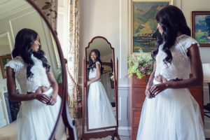 A woman in a white lace top and long tulle skirt smiles whilst looking at her reflection in a tall mirror in a softly lit, elegant room with floral decor and framed paintings.