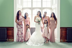 A bride in a white gown stands with five bridesmaids and two young flower girls in pink dresses, posing and smiling in front of a large, arched window inside an elegant room.
