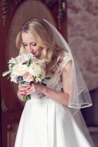 A bride in a white dress and veil holds a bunch of white and pale pink flowers, looking down and smiling softly.