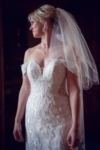 A bride in an off-the-shoulder, lace-embroidered wedding dress and veil stands in soft lighting, looking to the side with a serene expression.