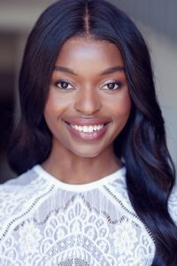 A woman with long, dark hair and smooth skin smiles warmly at the camera. She is wearing a white, lacy top and the background is softly blurred, keeping the focus on her face.