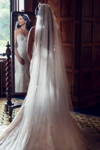 A bride in a lace wedding dress and long veil stands with her back to the camera, looking at her reflection in a tall mirror inside a wood-panelled room.
