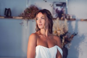 A woman in an off-the-shoulder white dress stands indoors, looking to the side. She has light brown hair styled loosely, and the background features shelves with flowers and decorative items.