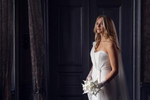 A woman in a white, off-the-shoulder wedding dress and lace gloves stands indoors holding a bouquet of white flowers, looking thoughtfully towards a window with sheer curtains. Dark panelled doors are in the background.
