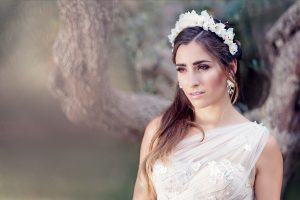 A woman in a white dress with floral details and a white flower crown stands outdoors near a tree, looking thoughtfully into the distance. The background is softly blurred.