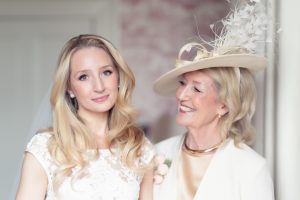 A bride in a white dress stands beside an older woman in a cream outfit and feathered hat. The woman smiles warmly at the bride, who looks calmly at the camera. Both have long blonde hair.
