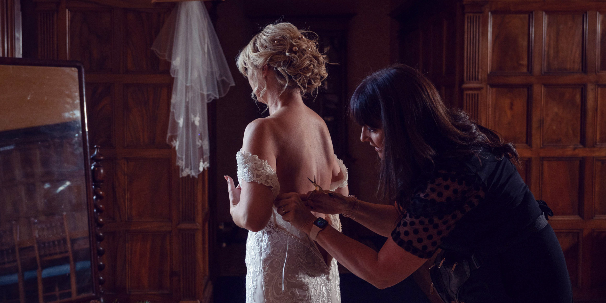 A woman helps a bride in a white lace wedding dress fasten the back of her gown in a dimly lit room with wooden panelling and a mirror reflecting part of the scene.