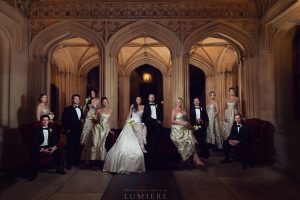 A wedding party poses in formal attire inside an ornate, arched hall. The bride and groom sit centre, surrounded by bridesmaids in gold dresses and groomsmen in black tuxedos, all looking at the camera.