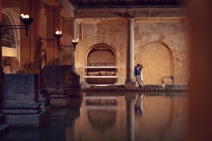 A couple stands close together on the edge of an ancient indoor Roman bath, surrounded by stone columns and softly lit torches reflecting on the still water.